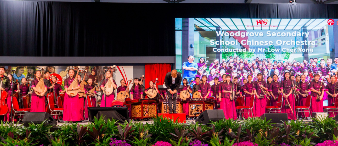 Woodgrove Secondary School Chinese Orchestra students in red perform on stage with a conductor bowing.